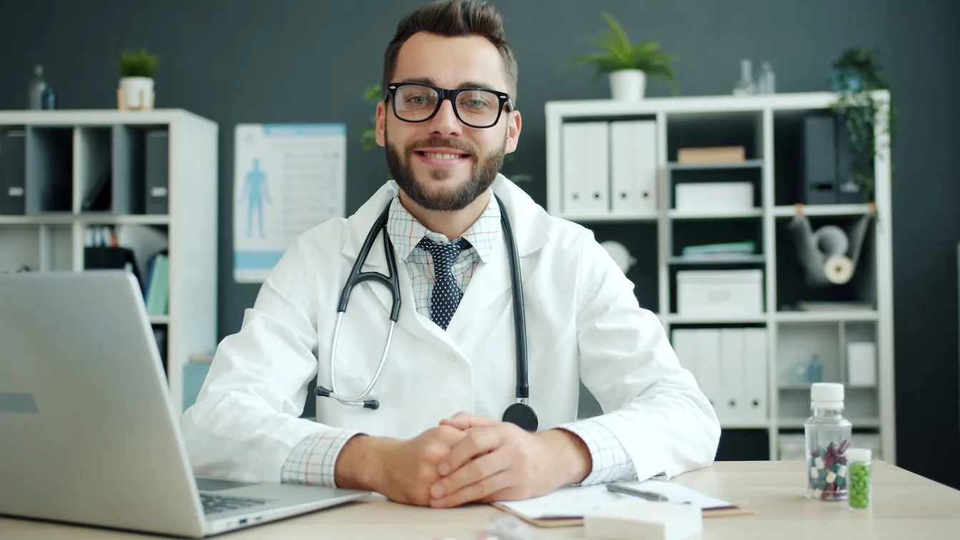 A doctor smiling at the camera while sitting at his desk infront of his computer 