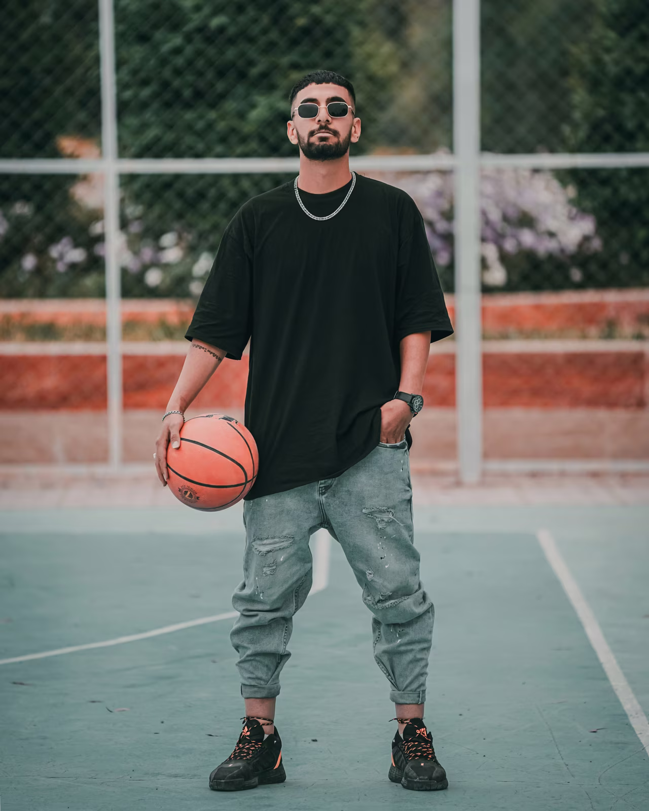 Man standing on an outdoor basketball court holding an orange basketball in his right hand, wearing sunglasses, a black oversized t-shirt, light blue ripped jeans, and black sneakers.