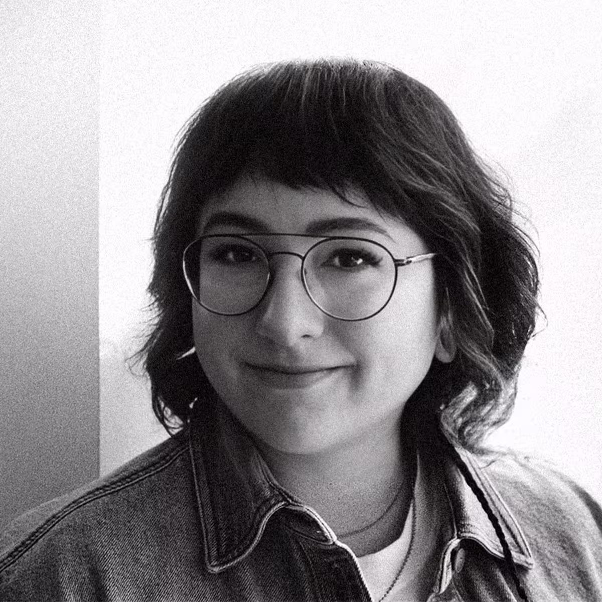 Smiling woman with brown hair tied back, wearing large brown glasses and a black top, in a modern indoor setting with blurred architectural background.