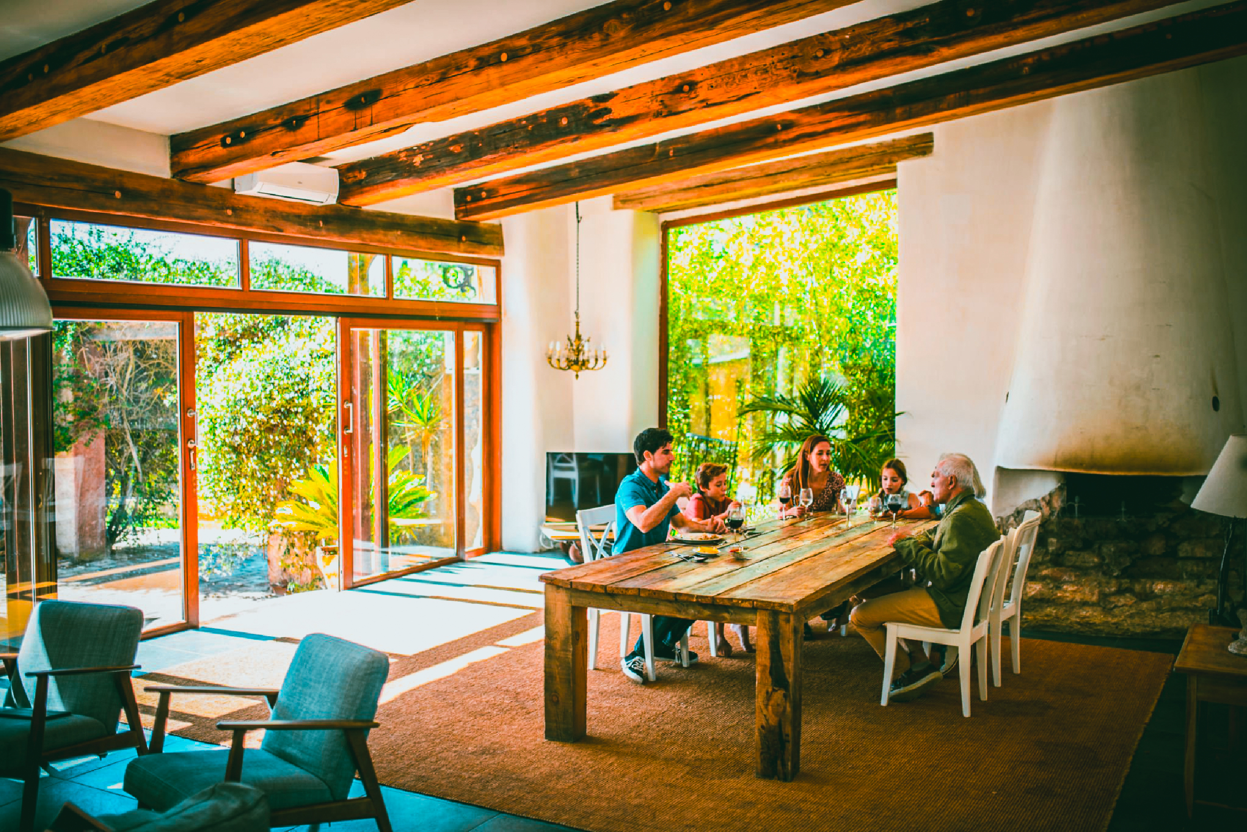 A family sits at a long wooden table in a sunlit open living room.