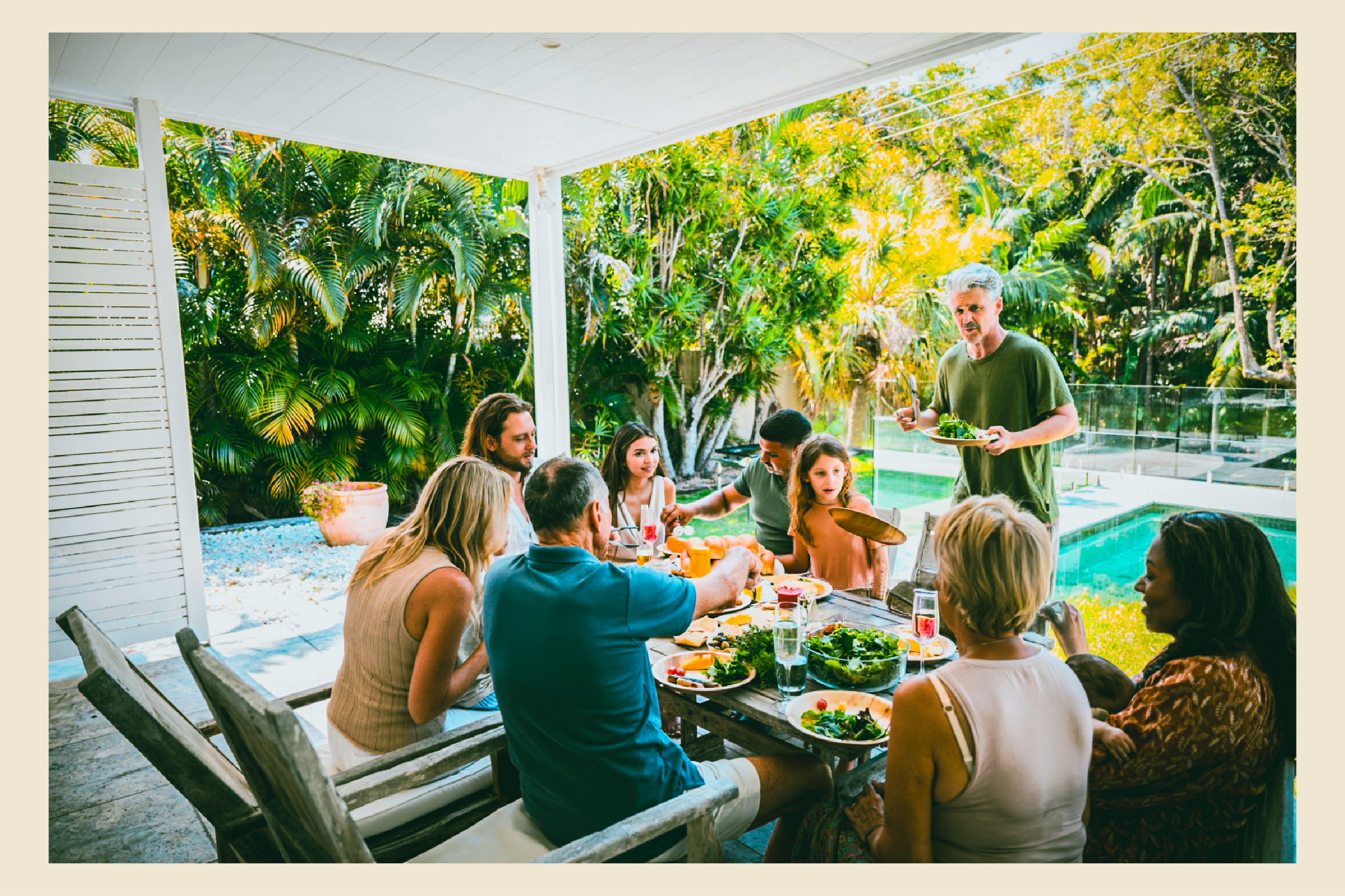Adults share a meal at an outdoor table beside a swimming pool.