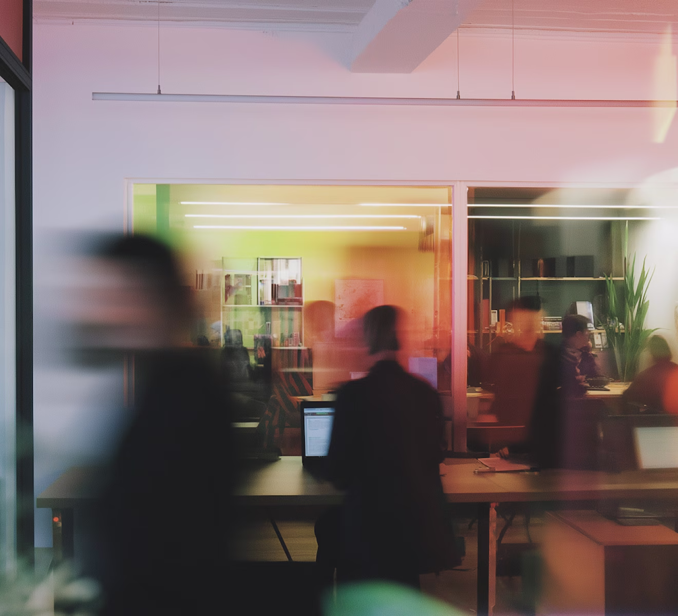 Blurred image of people working in an office with desks, computers, and bookshelves visible through glass walls.