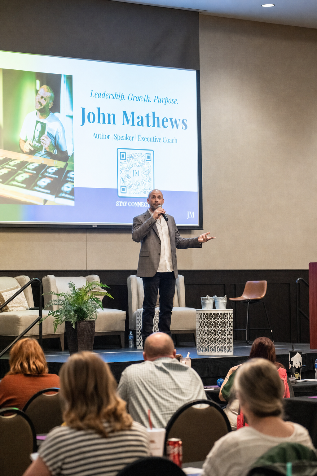 John Mathews speaking into a microphone with an open-hand gesture, with seated audience members in the foreground