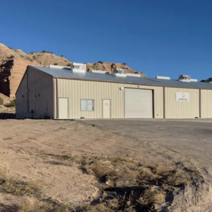 Large beige metal industrial building with a white garage door and small windows, set against a clear blue sky and rocky hills background.