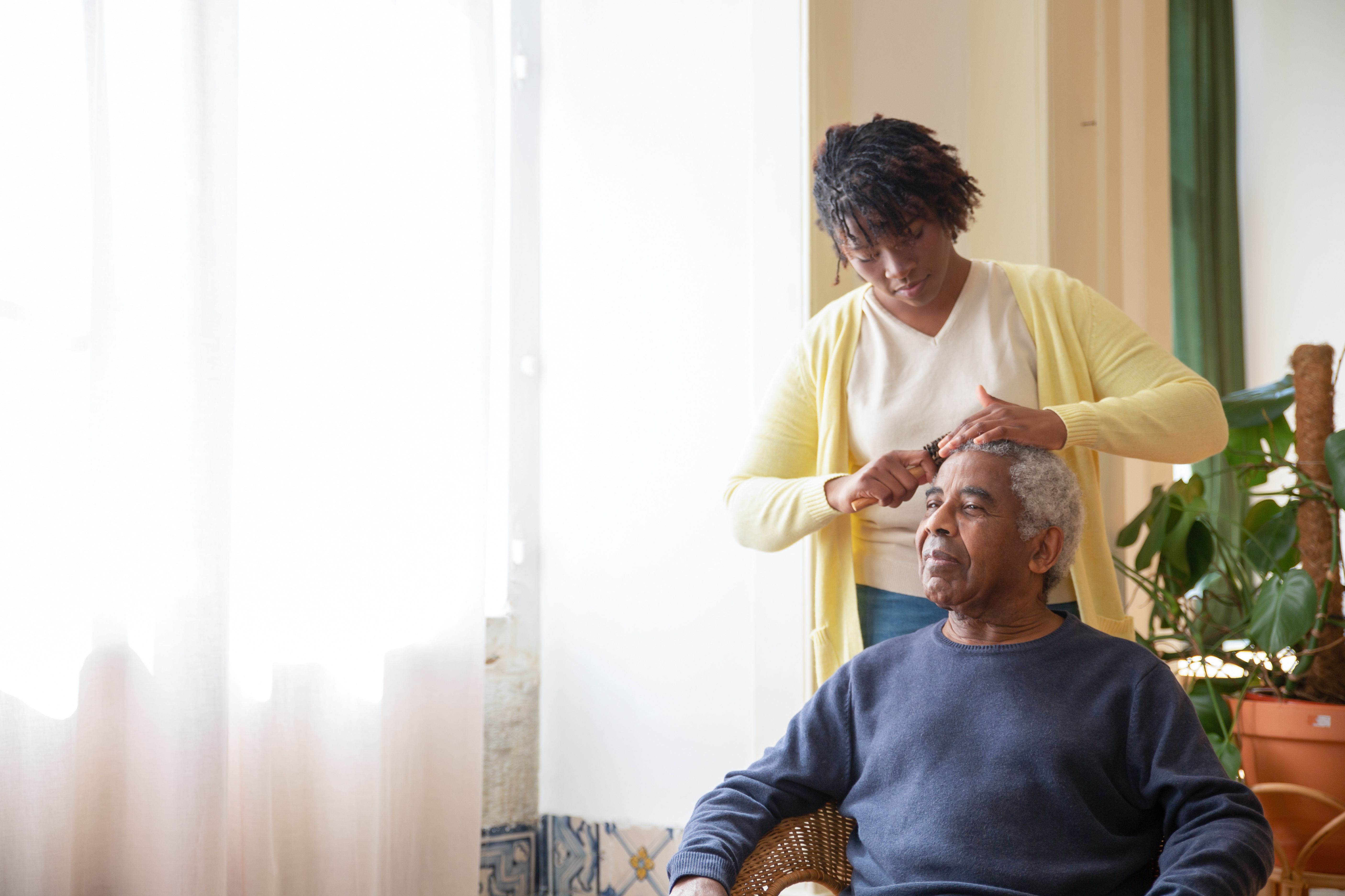 Home health caregiver gently combing an older man's hair beside a sunlit window in his living room