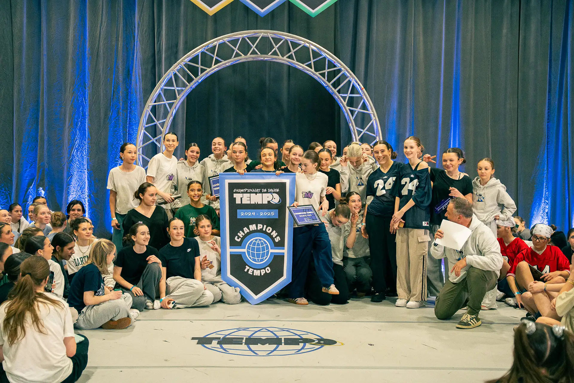 Une équipe championne pose avec la bannière Champions Tempo devant l'arche du plancher, au Championnat de danse Tempo 2025
