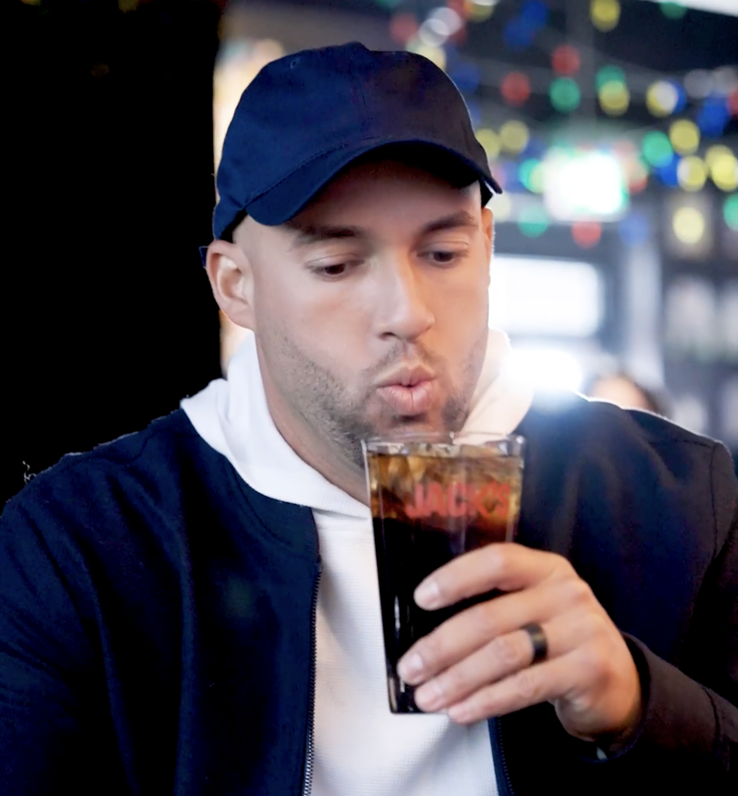 George Springer sipping a dark beverage in a navy cap and jacket at a bar with colorful bokeh lights