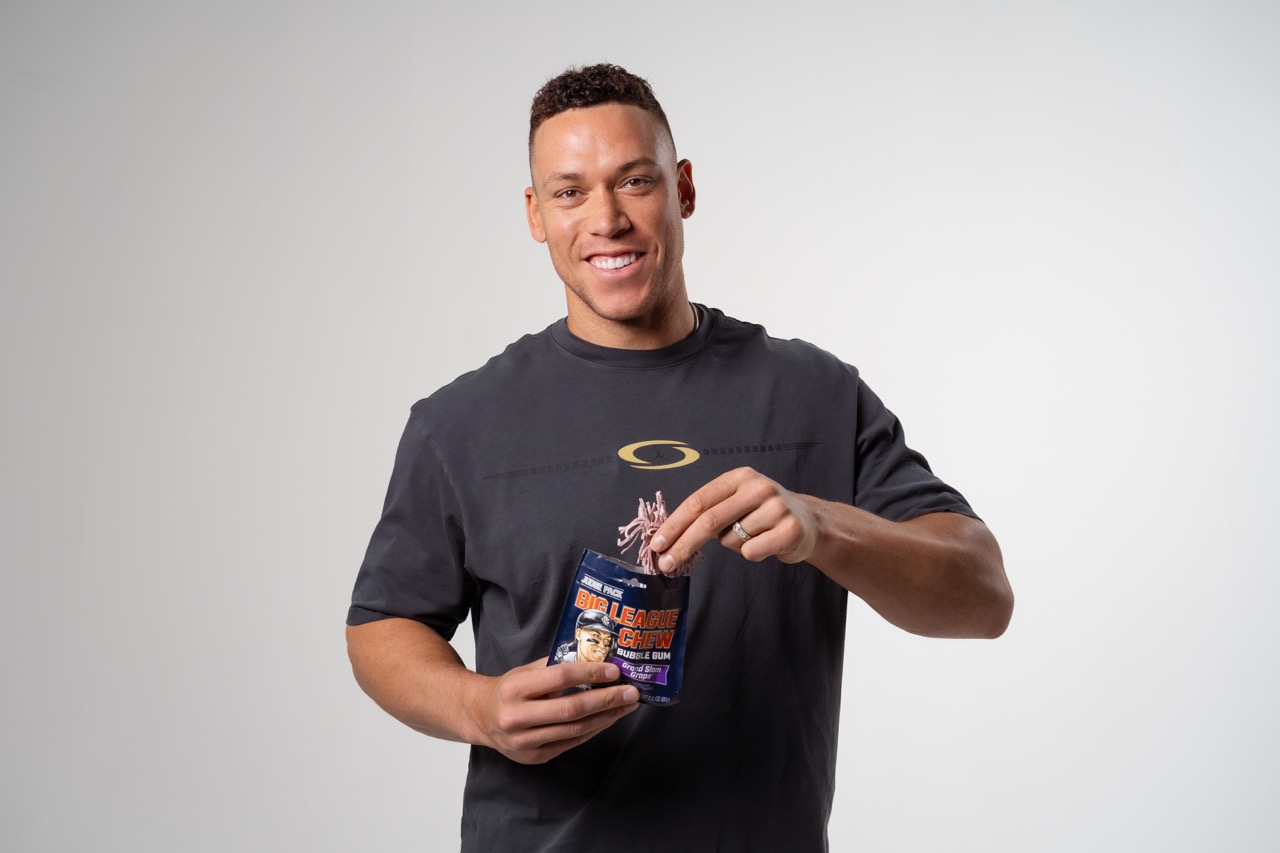 Aaron Judge smiling while holding a bag of Big League Chew gum, wearing a black t-shirt