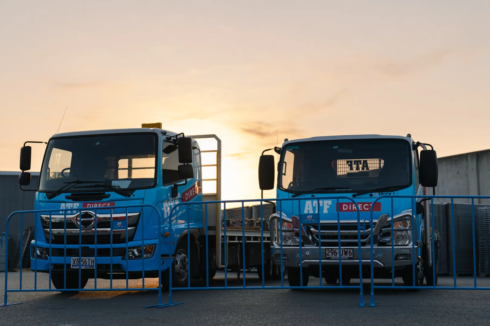 Two blue ATF Direct trucks parked behind blue metal barricades at sunset.