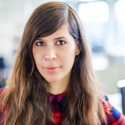 Close-up portrait of a woman with long brown hair and brown eyes, wearing a red and blue plaid shirt, with a softly blurred office background.