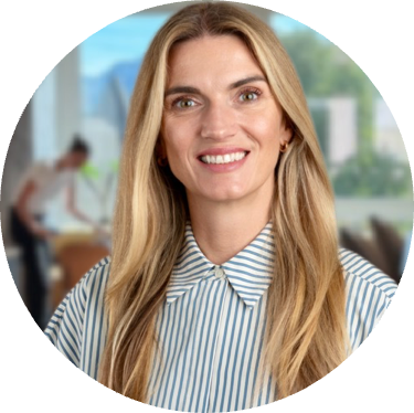 Smiling woman with long blonde hair wearing a blue and white striped collared shirt, with a blurred office background.