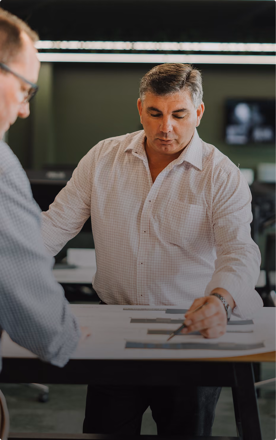 Two men in an office collaborating over printed charts or plans on a black table, one pointing with a pen while the other looks on attentively.