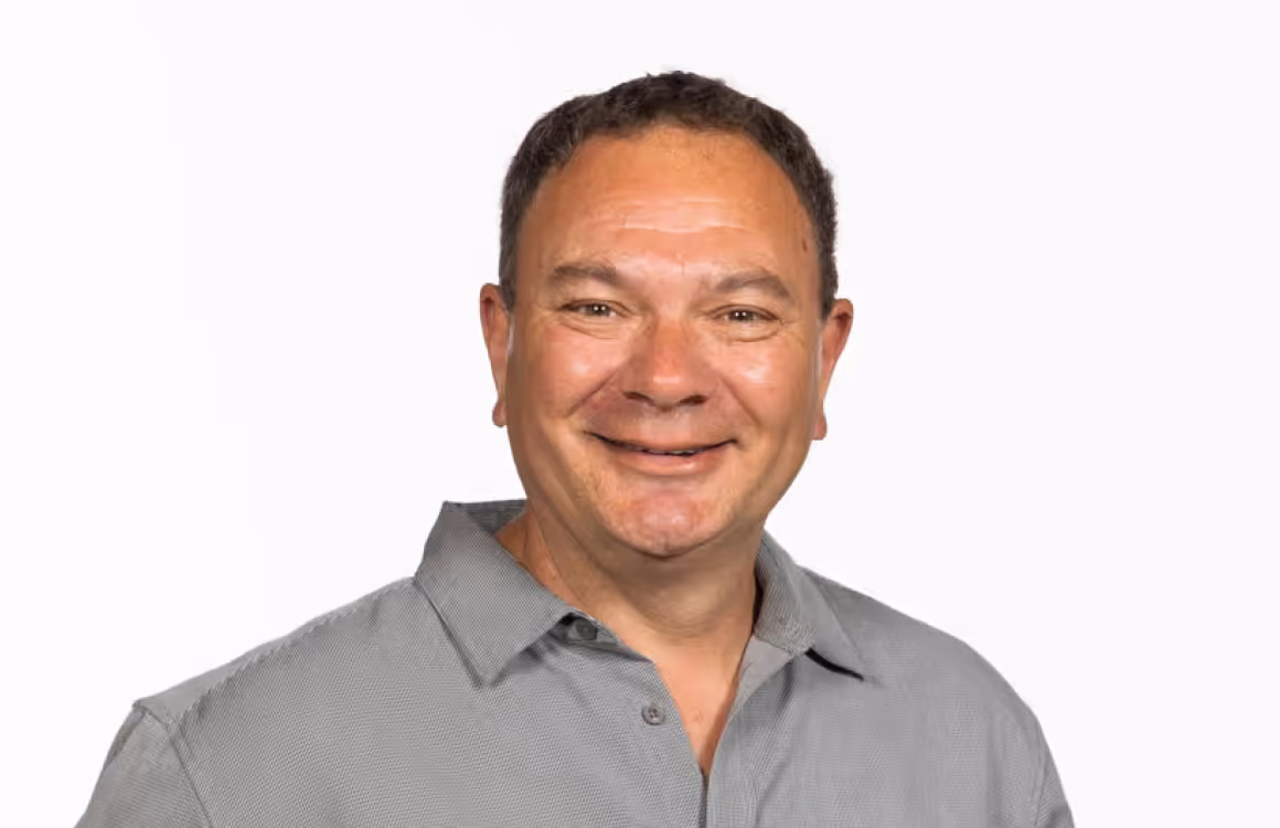 Smiling man with short dark hair wearing a gray button-up shirt against a white background.