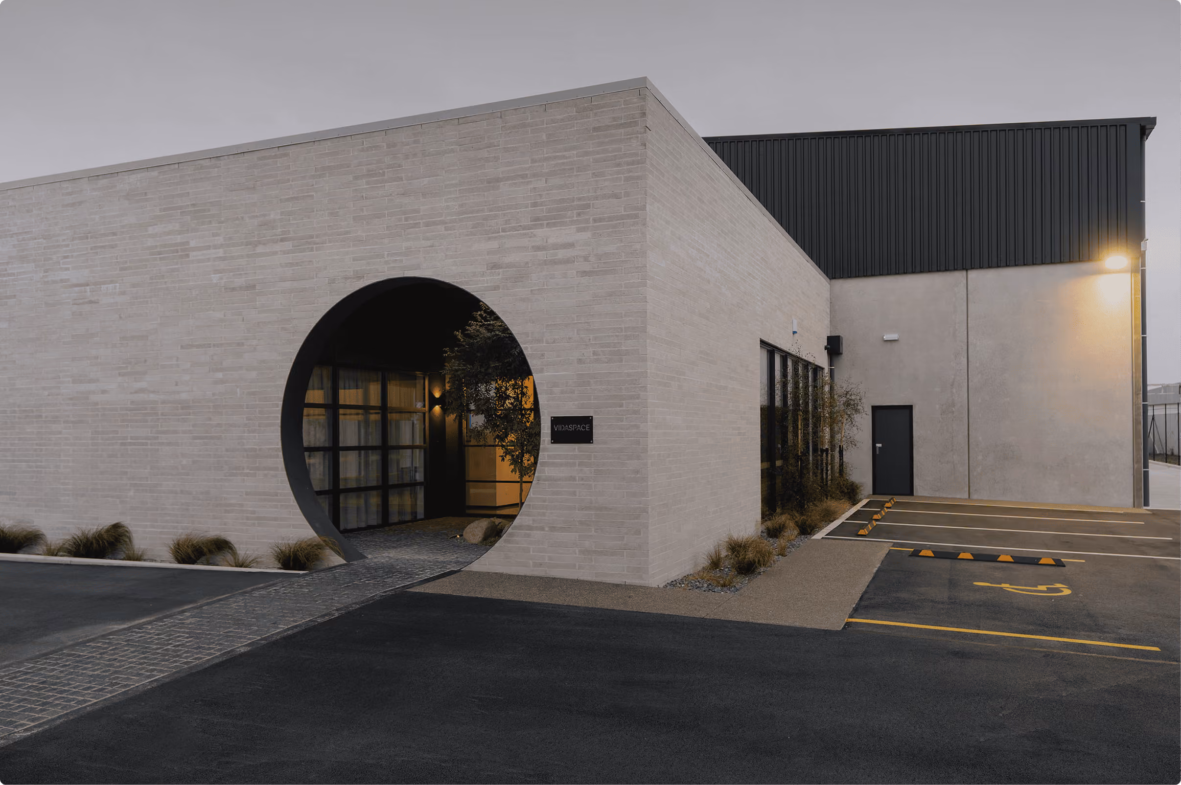 Modern building with a large circular entryway framed in light-colored bricks, leading to an illuminated interior space with glass windows and a sign reading 'WORKSPACE', adjacent to a parking lot with empty spaces and a handicapped parking spot.