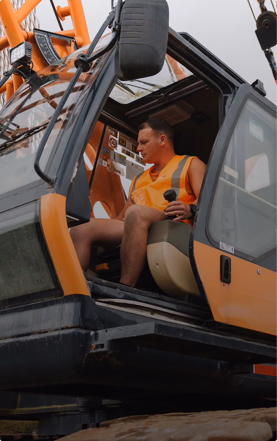 Construction worker in an orange safety vest operating controls inside the cabin of an orange heavy machinery crane.
