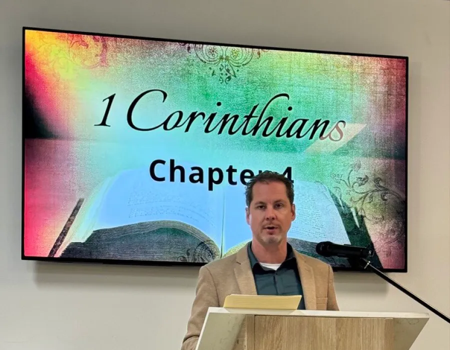A man in a tan blazer and dark shirt stands behind a wooden podium with a microphone, speaking in front of a large screen displaying '1 Corinthians Chapter 4' with an open Bible image in the background.