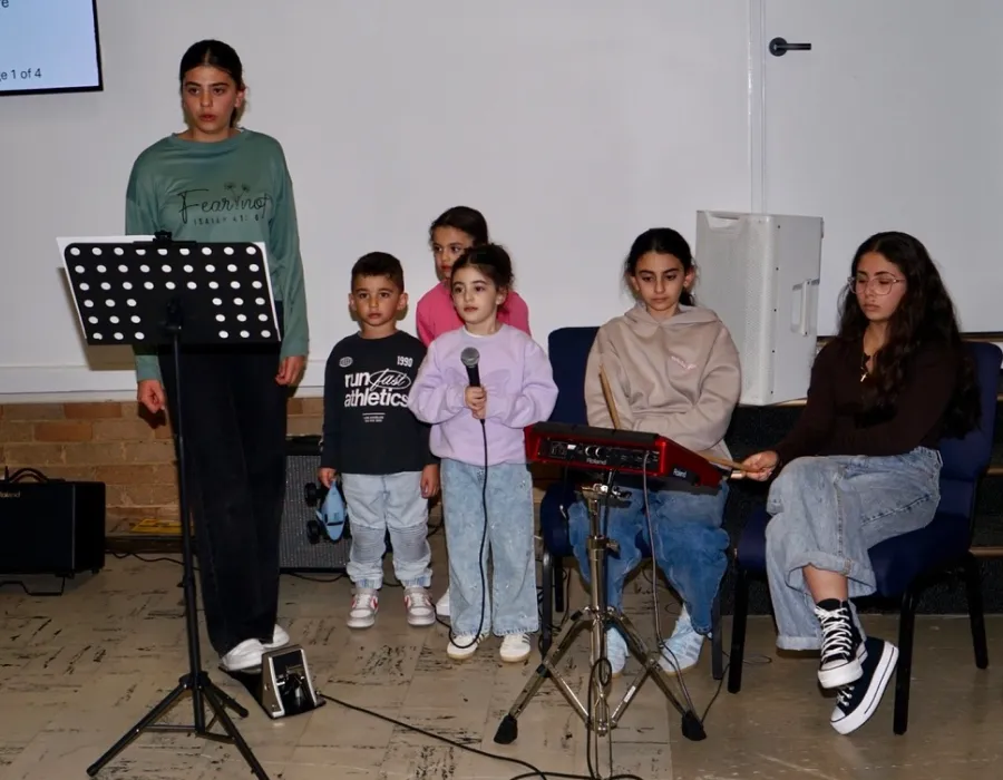 Six children indoors: one standing at a music stand wearing a green sweatshirt, two girls and one boy in the middle with one girl holding a microphone, and two seated girls on the right, one playing a small electronic keyboard, the other holding drumsticks.