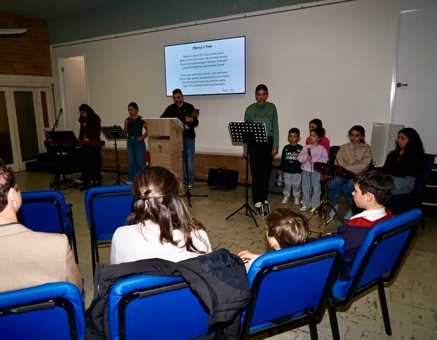 Group of people, including children and adults, performing music and singing indoors with a screen displaying song lyrics titled "Mercy's Tree" in the background, while an audience sits in blue chairs.