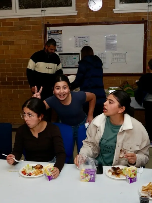 Three young people sitting and eating at a table with plates of food and juice boxes, while a girl standing behind them smiles and makes a peace sign over another's head in a casual indoor setting.