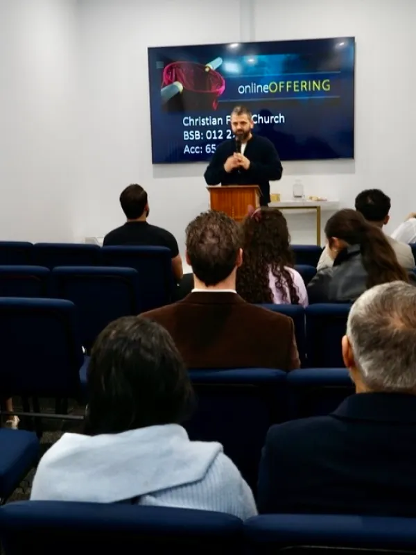 Man speaking at a podium to an audience seated in blue chairs in a small room, with a screen behind him displaying 'online OFFERING' and Christian Faith Church banking details.