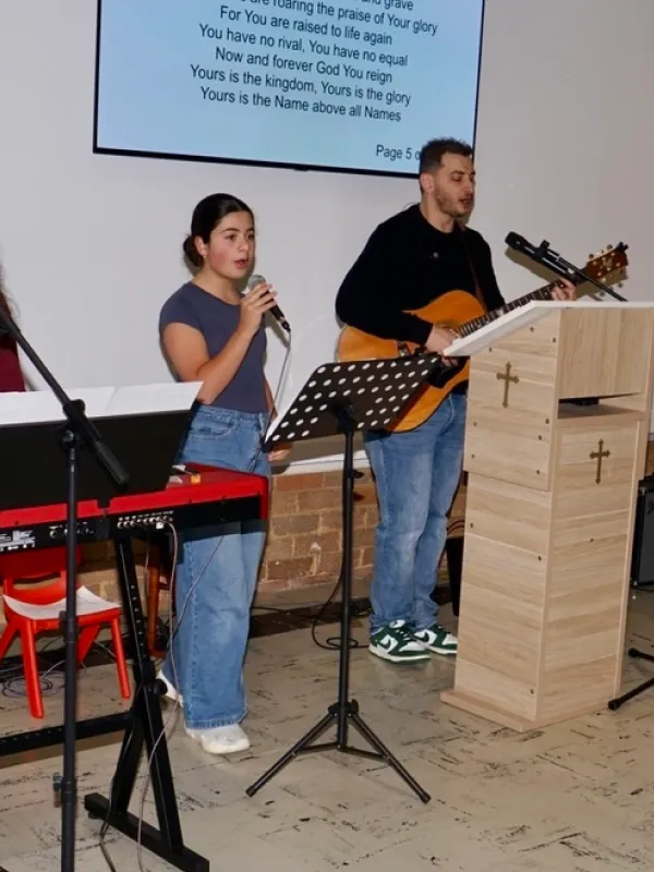 A young woman holding a microphone and singing next to a man playing an acoustic guitar in a church setting with a wooden podium featuring crosses and a screen displaying worship lyrics behind them.