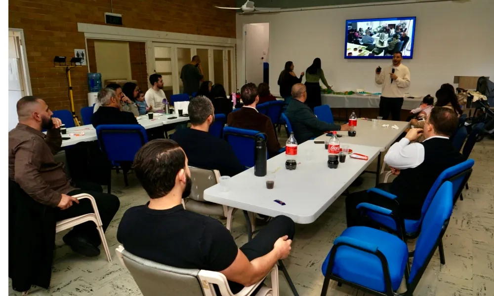 A group of people seated around tables in a meeting room, attentively watching a man standing and speaking near a TV screen displaying a video conference or presentation.