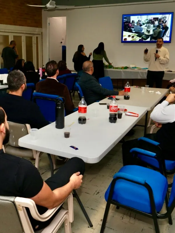 A group of people seated around tables with bottles of Coca-Cola while a man stands and speaks holding a microphone in a meeting or presentation room with a wall-mounted screen displaying a similar scene.