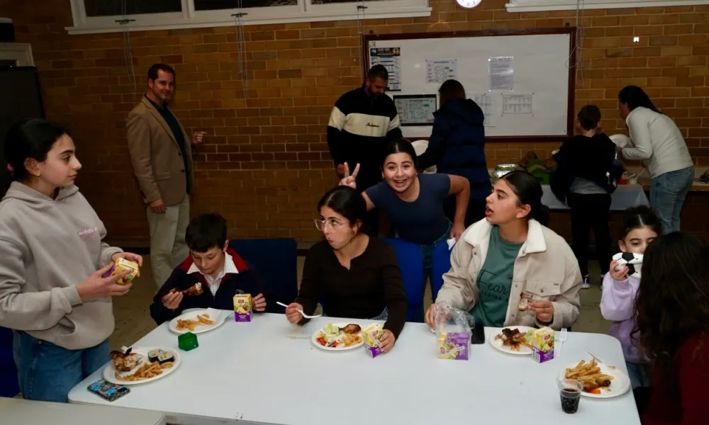 Group of children and a few adults in a cafeteria or community room eating meals with plates of food, juice boxes, and drinks on a white table, with a brick wall and whiteboard in the background.