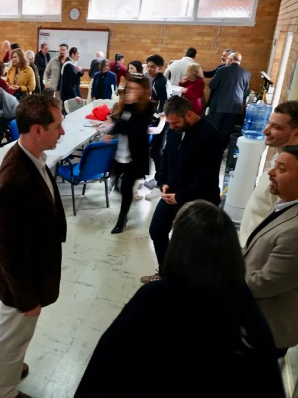 A group of people gathered in a room with brick walls and windows, some standing and talking, others walking past tables with blue chairs, a water dispenser visible in the background.
