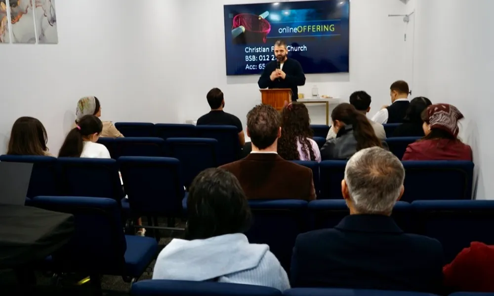 A man speaking at a podium in a small church or meeting room with an audience seated in rows facing him, and a screen behind displays text about online offering for Christian Faith Church with bank details.