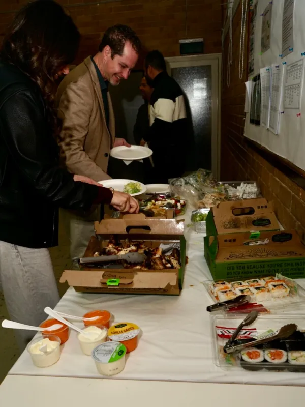 People serving themselves food at a buffet table with cardboard pizza boxes, sushi trays, salads, and various sauces in small containers with spoons.