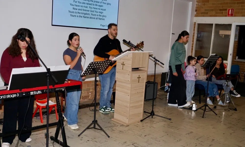 A group of seven people performing in a church setting; one woman plays a keyboard, a man plays an acoustic guitar nearby, and two women sing into microphones while others stand or sit holding microphones. A wooden pulpit with crosses is centered, and a screen behind them displays song lyrics.