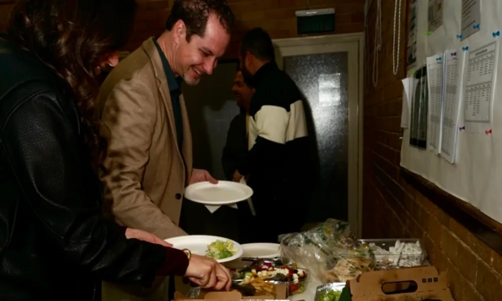 Two people serving themselves food from a buffet table with plates, salad, and various dishes in a dimly lit room.
