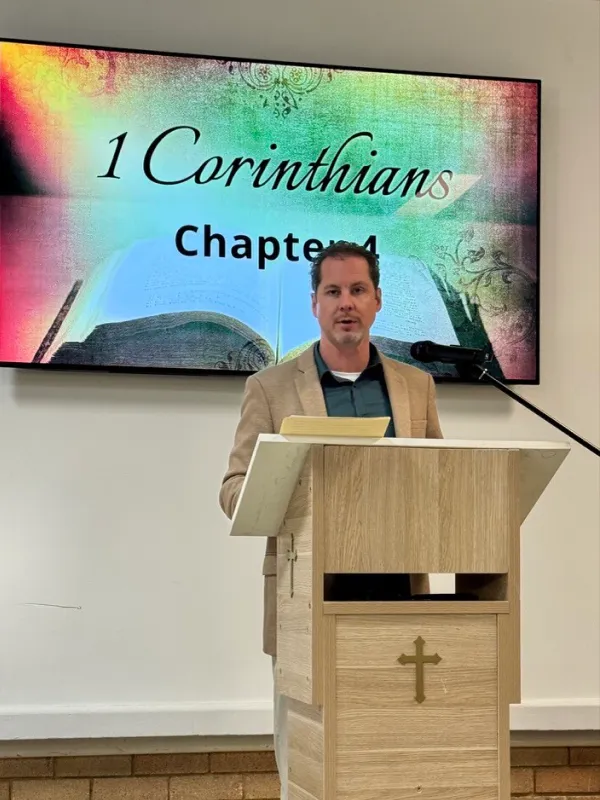 Man standing behind a wooden pulpit with a cross symbol, speaking in front of a screen displaying '1 Corinthians Chapter 4' with an open Bible background.