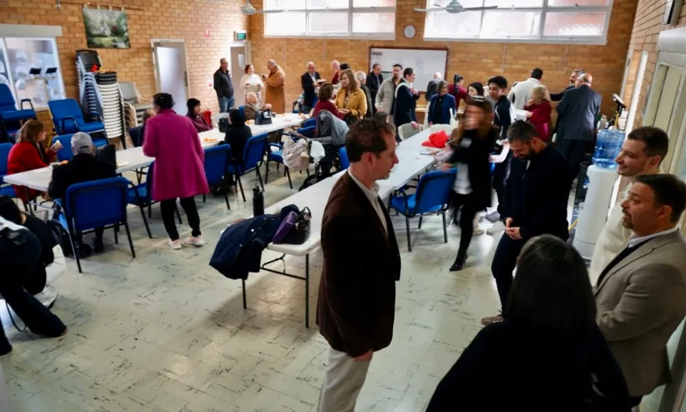 Indoor community gathering with multiple people engaged in conversation around tables and standing, in a room with brick walls and large windows.