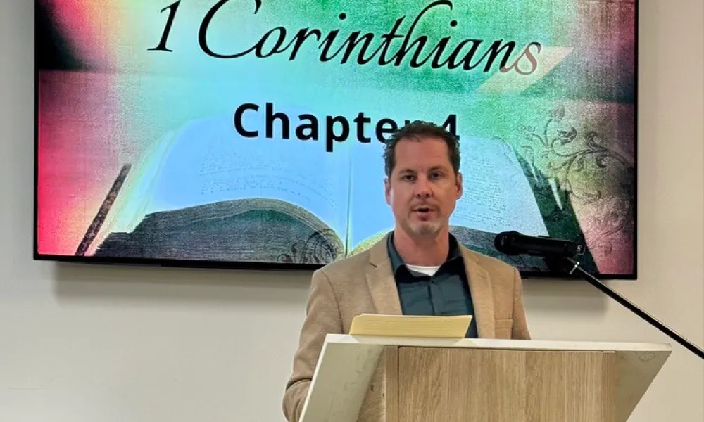 A man stands at a wooden podium speaking or presenting with a microphone to his right, in front of a screen displaying the text '1 Corinthians Chapter 4' and an image of an open Bible.