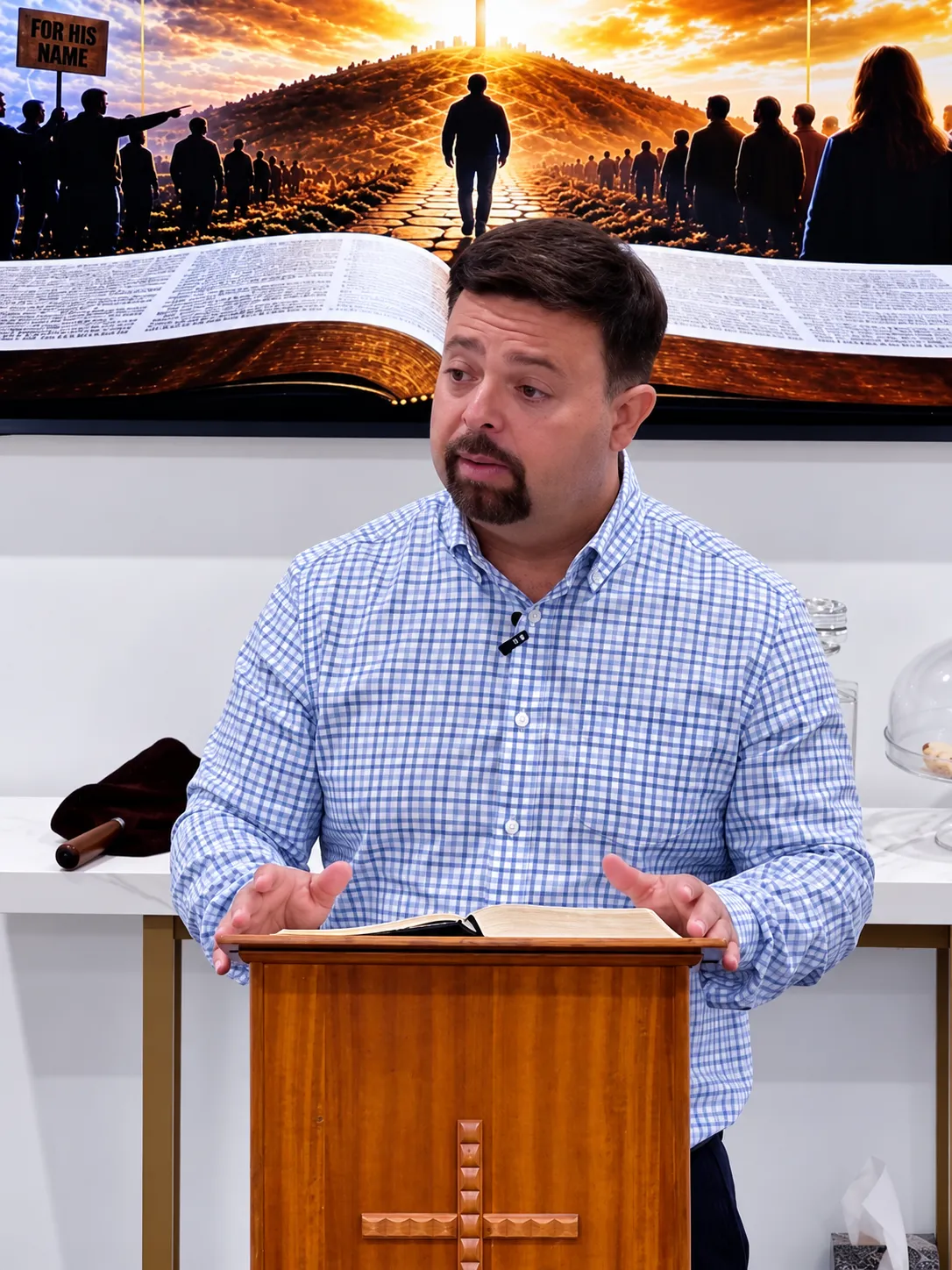 A man in a blue and white checkered shirt stands behind a wooden lectern with a cross, speaking with an open book in front of him and a large artwork of a glowing path and people behind him.