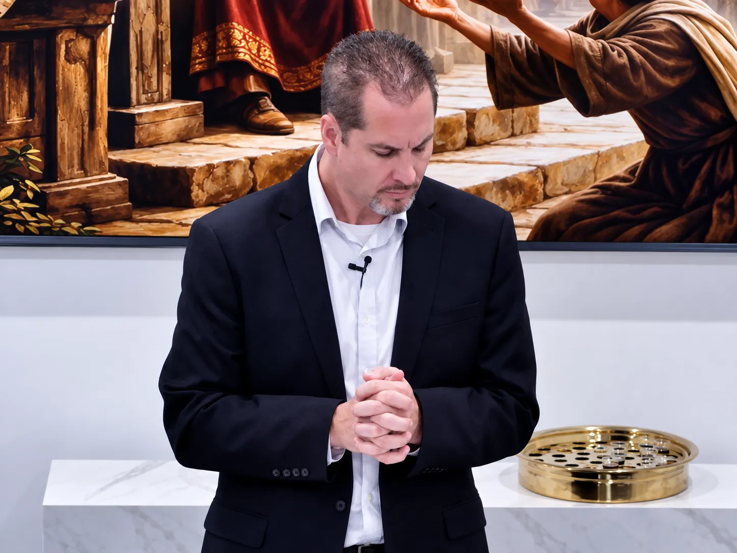 A man with short hair and a goatee, wearing a black suit jacket over a white shirt, stands with his hands clasped in front of him and looks downward in prayer or contemplation, with a religious painting and a gold communion tray with small cups behind him.