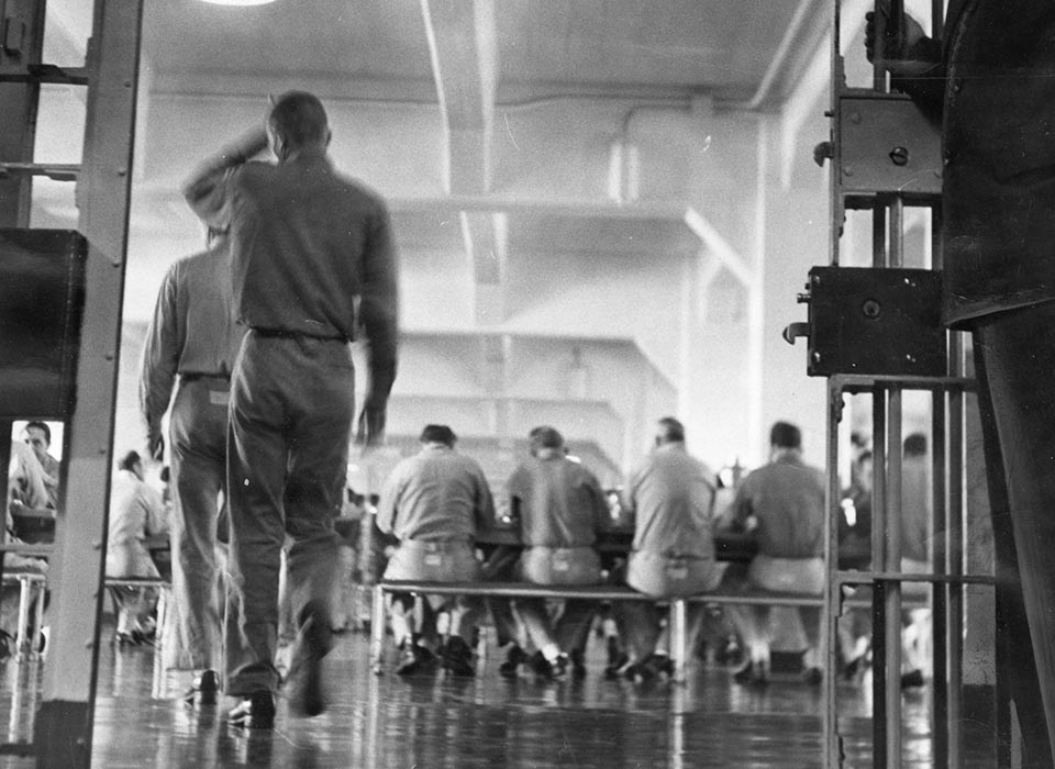Archival black-and-white photograph of men seated at tables in the Alcatraz dining hall, with a uniformed officer in the foreground.