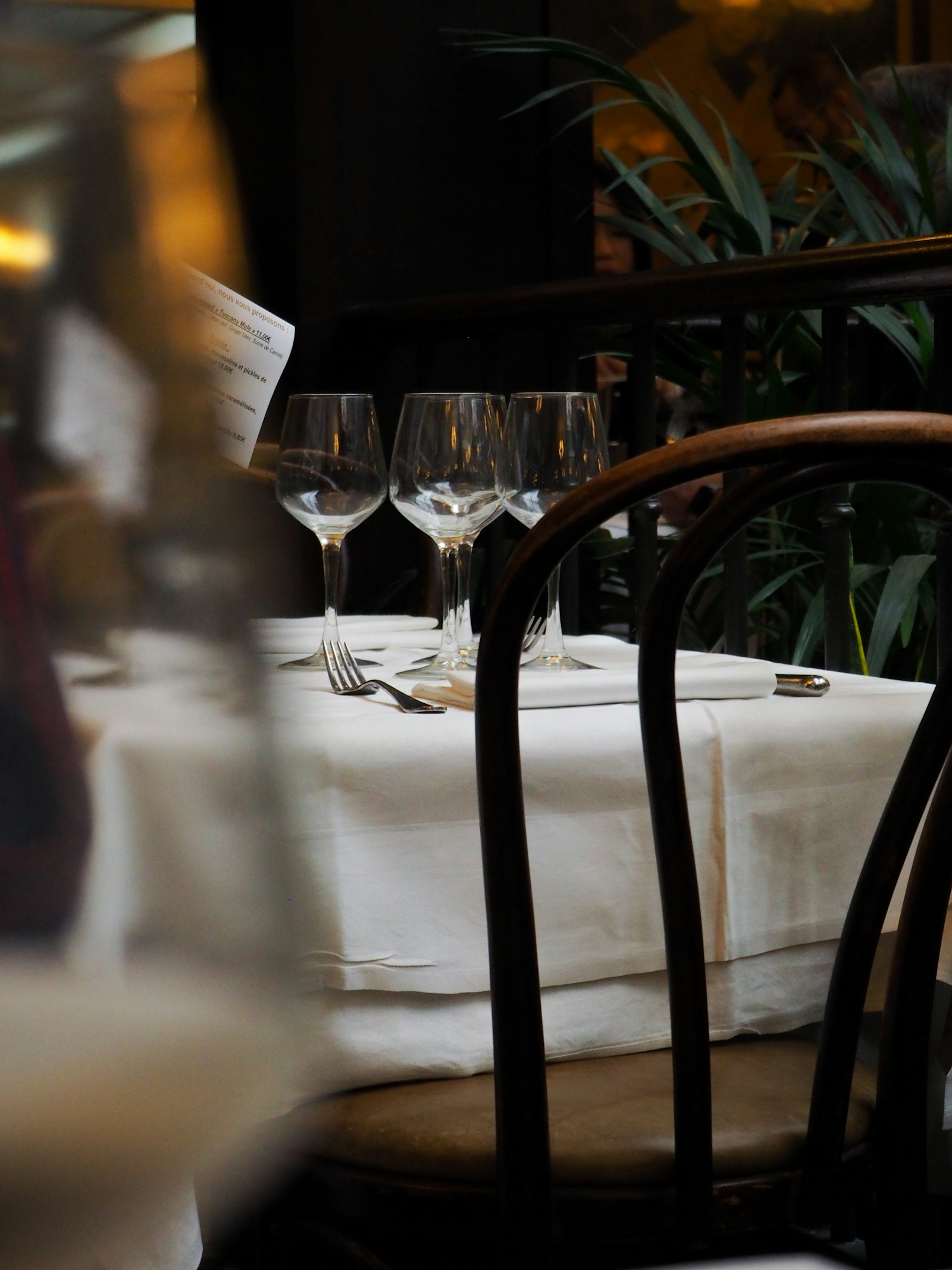 Empty wine glasses and cutlery on a white tablecloth, seen past the back of a dining chair.