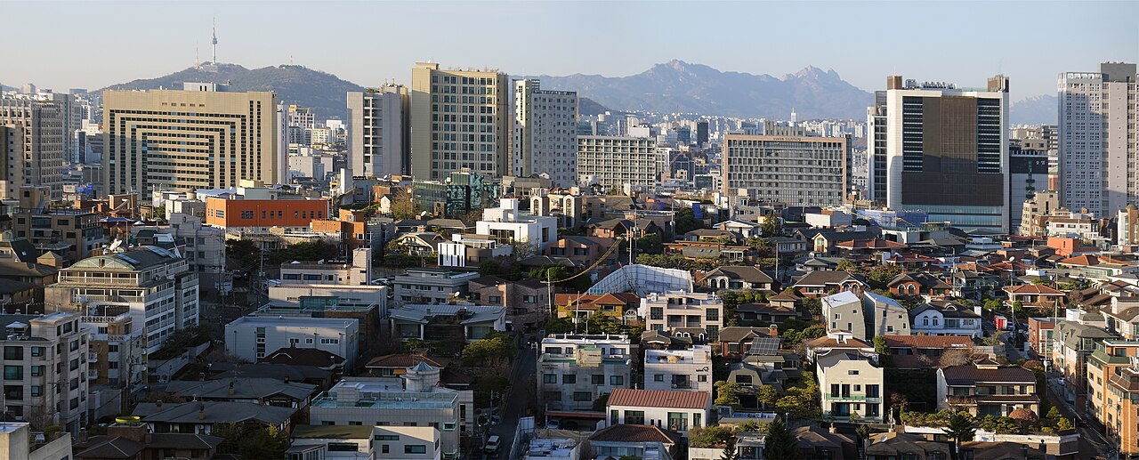 A panoramic view over the low-rise buildings and office towers of Gangnam-gu from Yeoksam, with hills beyond.