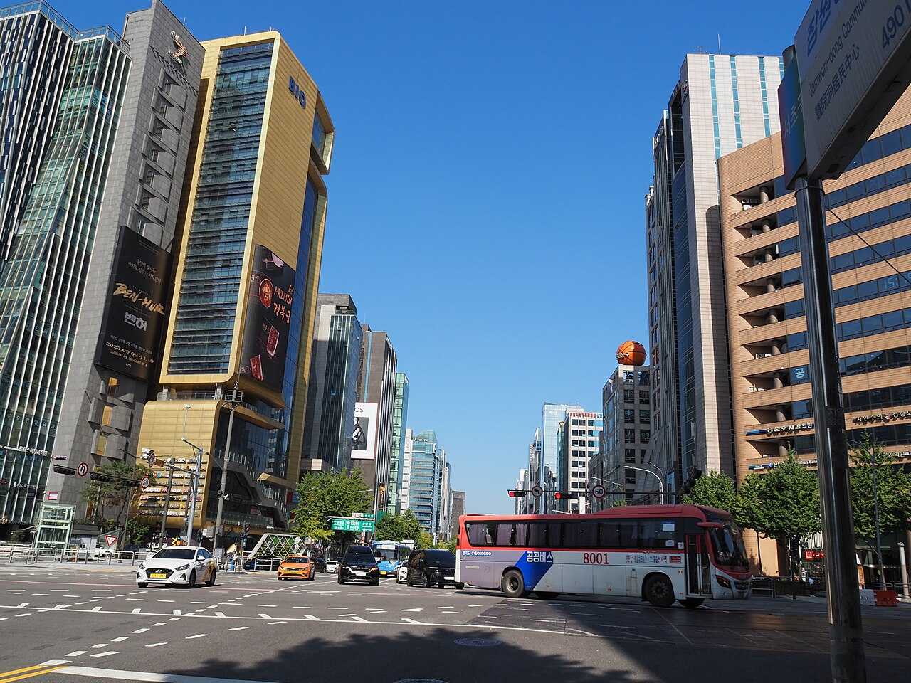 A street view in Sinsa-dong with buses, traffic and buildings of different heights beneath a blue sky.