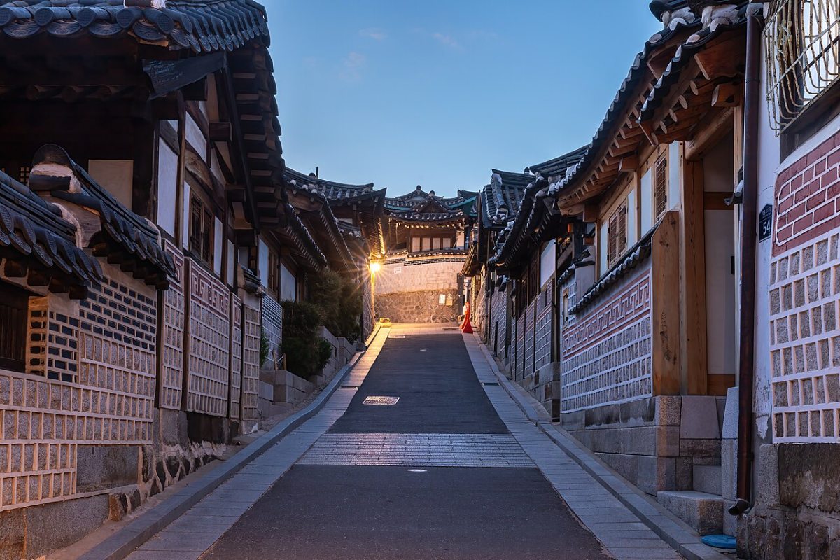 A sloping Bukchon street lined by brick walls, timber doors and tiled roofs at blue hour in June 2024.