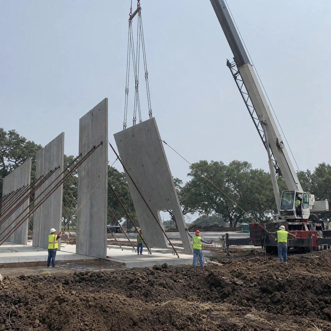 Mobile crane lifting a concrete tilt-wall panel upright on a Houston-area commercial construction site with braced panels behind