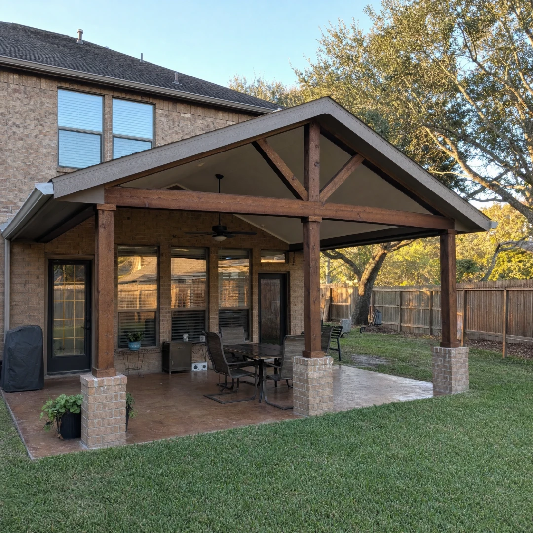 Finished gabled patio cover with cedar-stained posts and a shingle roof attached to the back of a two-story brick home