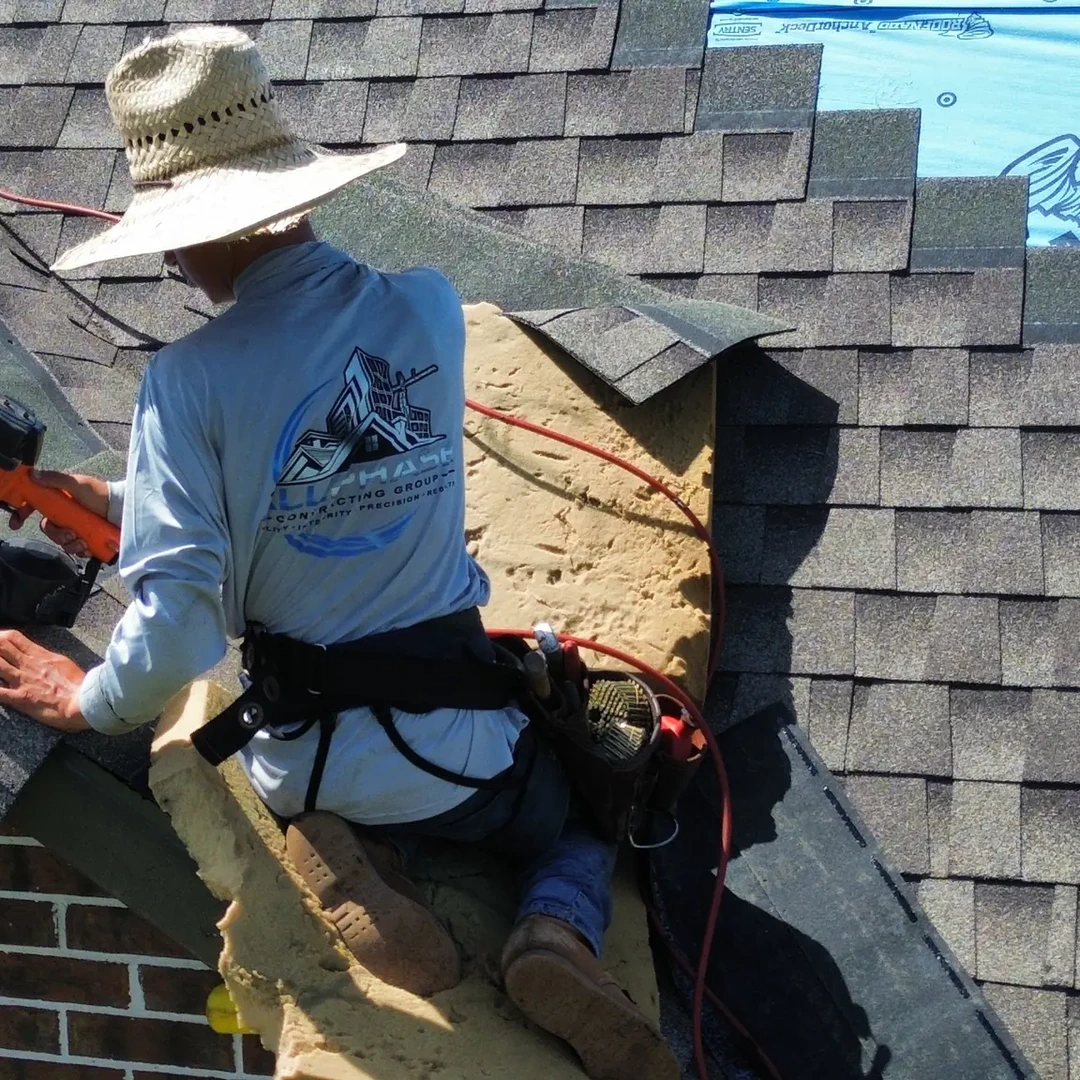 Roofer in a wide-brim hat nailing architectural asphalt shingles into place near the ridge of a residential roof