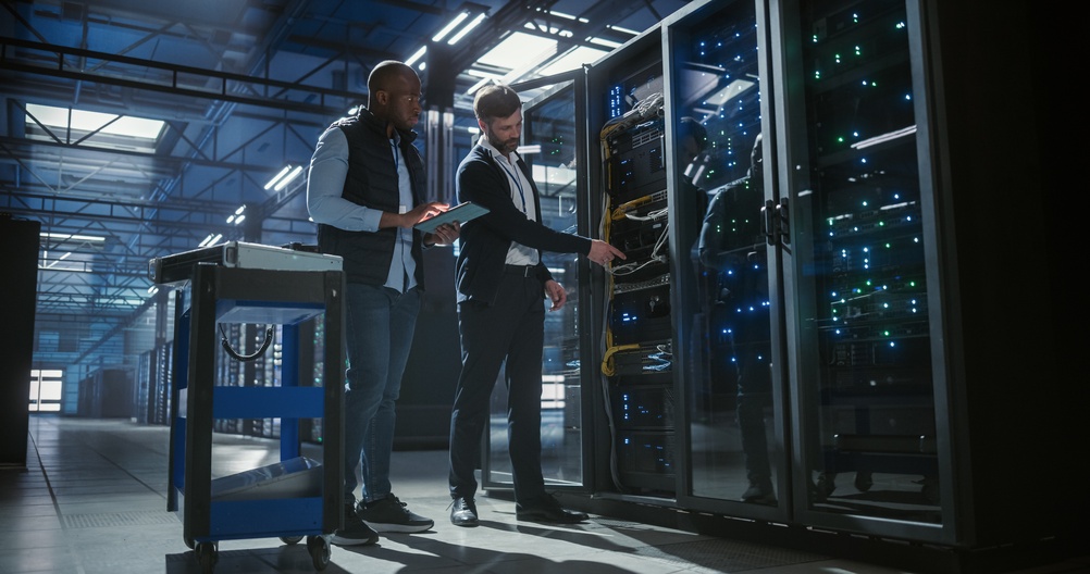 Two IT professionals in a data center examining server racks; one man holding a tablet, the other pointing at equipment inside a glass server cabinet with blinking blue and green lights.