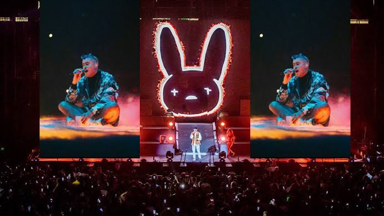 Singer Bad Bunny performs on stage with two large screens showing him singing while sitting cross-legged and a large fiery bunny face logo lit up behind him, with a crowd holding up phones in front.