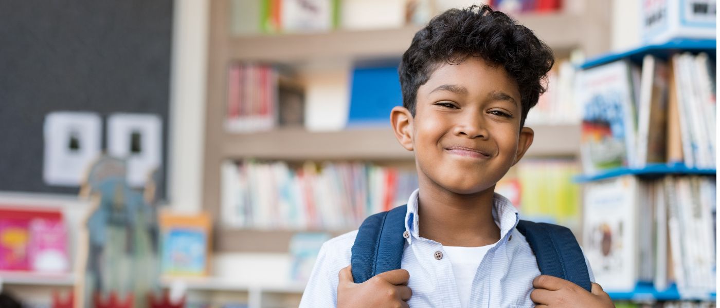 Smiling young boy with curly hair in a light blue shirt holding the straps of his blue backpack in a library.