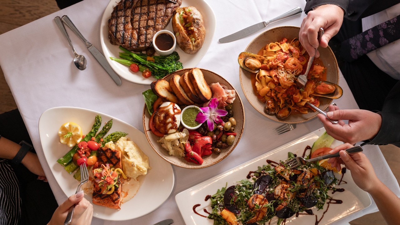 Top view of a dining table with four plates of gourmet food including a grilled steak with baked potato and vegetables, a seafood pasta, a plate of grilled salmon with asparagus, mashed potatoes, and tomatoes, and a salad with grilled shrimp and balsamic drizzle, with three people holding forks and knives ready to eat.